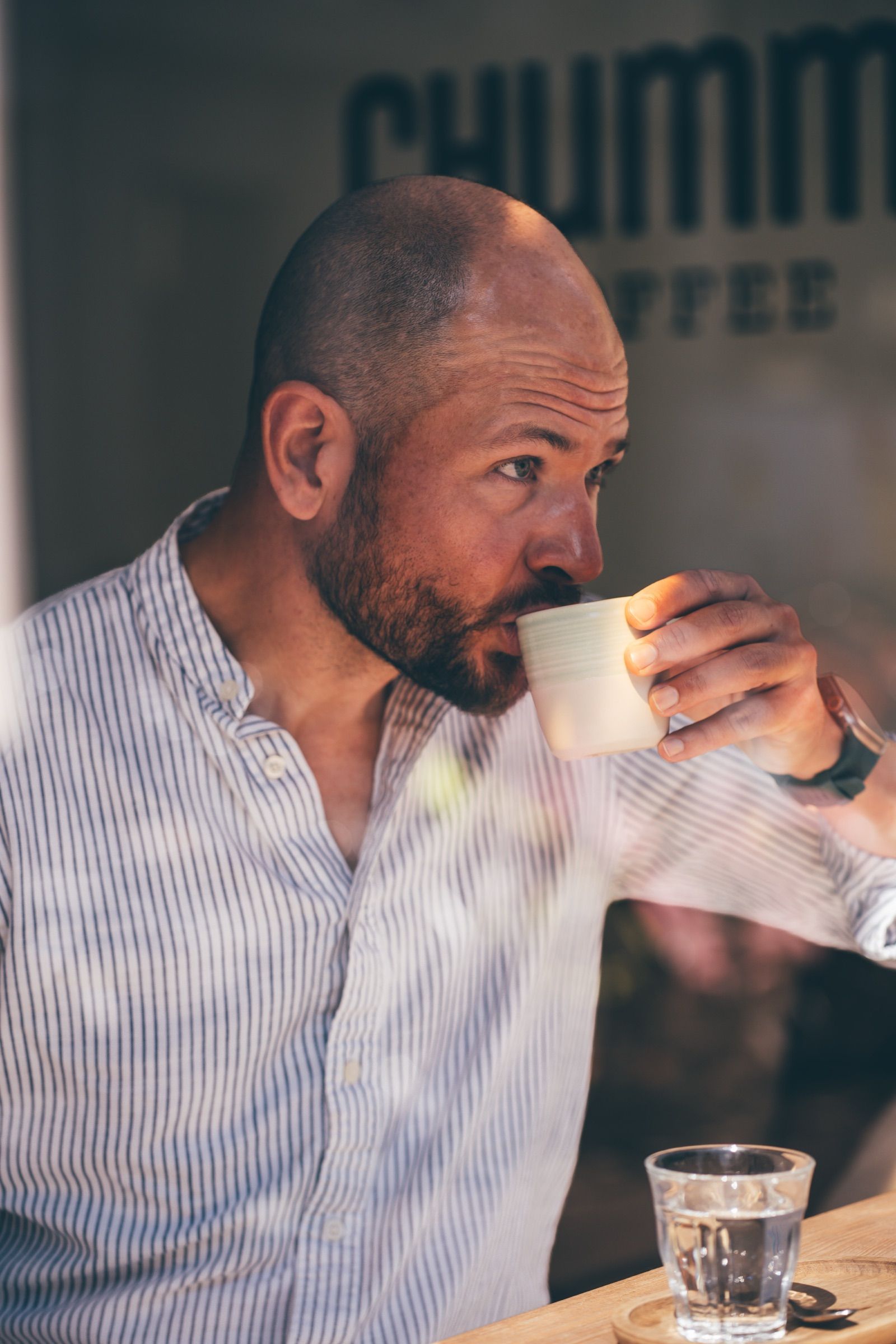 Bald bearded man in a blue-striped shirt sipping from a ceramic cup at a sunlit café table, a coffee-shop sign softly out of focus behind him